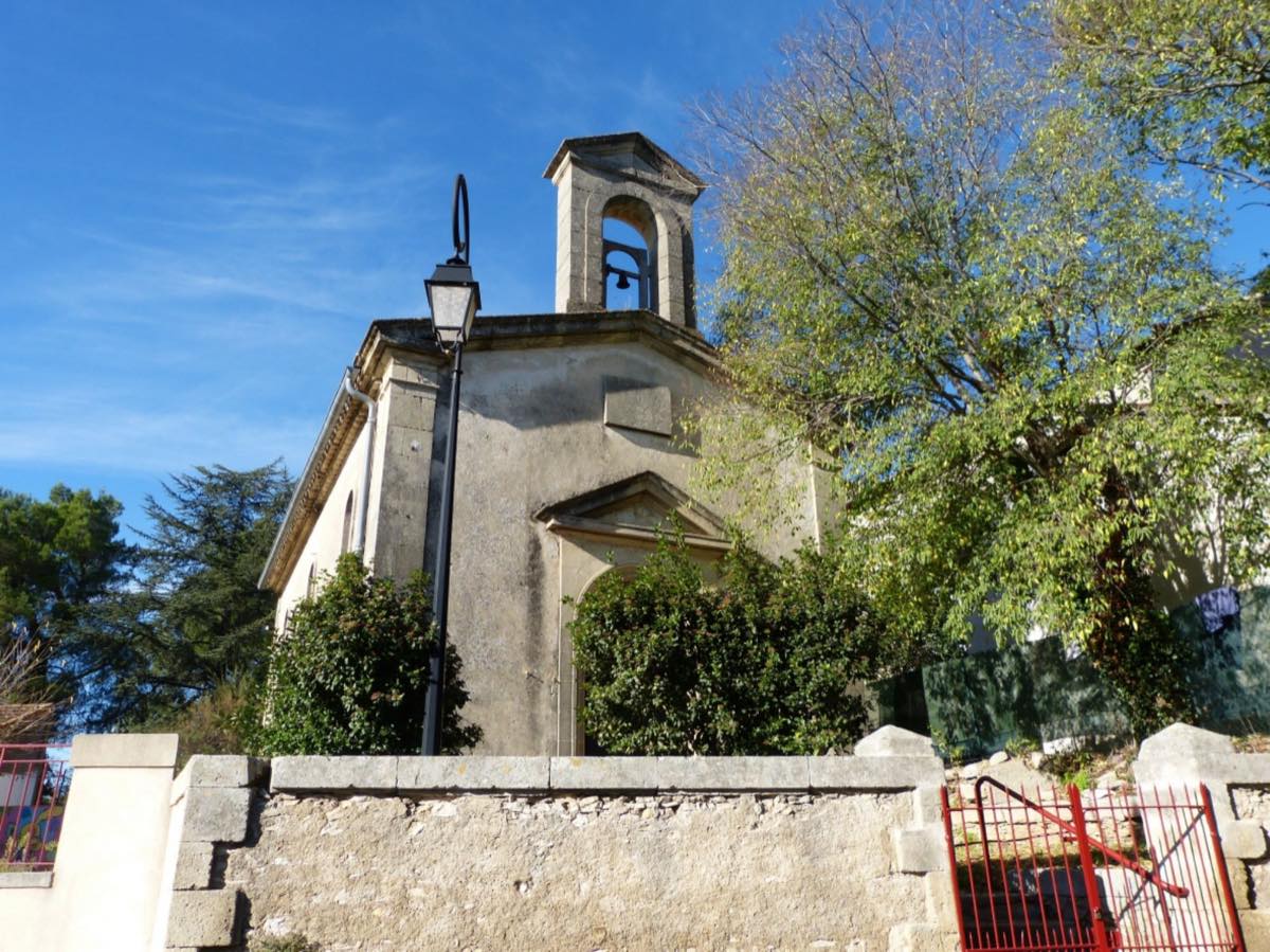 L'église de Lecques vue depuis la rue, avec son clocher-mur surmonté d'une cloche, derrière un mur de pierre sèche.
