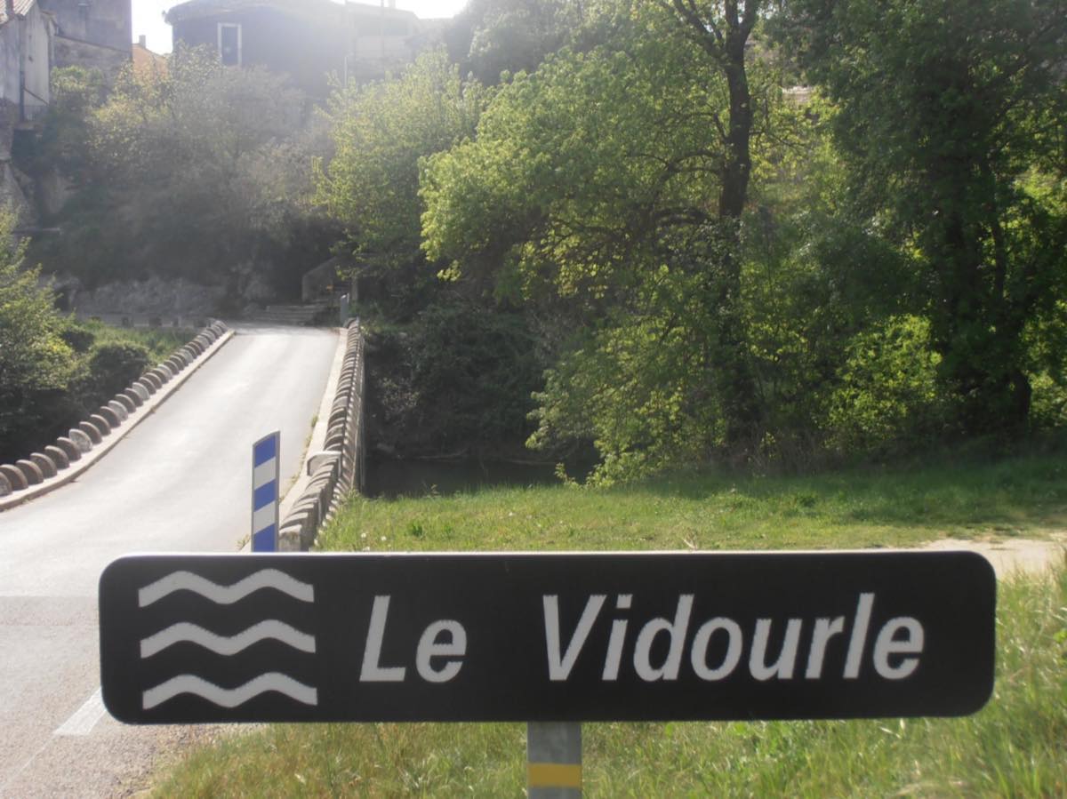 Le panneau routier « Le Vidourle » planté au bord de la chaussée, juste avant le pont qui franchit la rivière.