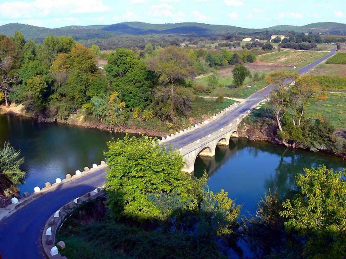Vue aérienne du pont de pierre à arches franchissant le Vidourle, entouré de végétation et des collines de garrigue.